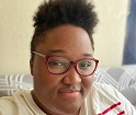 A woman looking at the camera, her natural hair short on the side and a lovely puff at the top. She has red-rimmed glasses and a gentle smile.